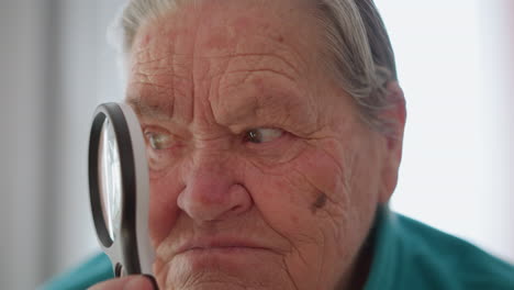 elderly woman in green jacket holding magnifying glass close to her face, staring through lens with serious expression, focused gaze, sitting indoors, natural light shining from behind
