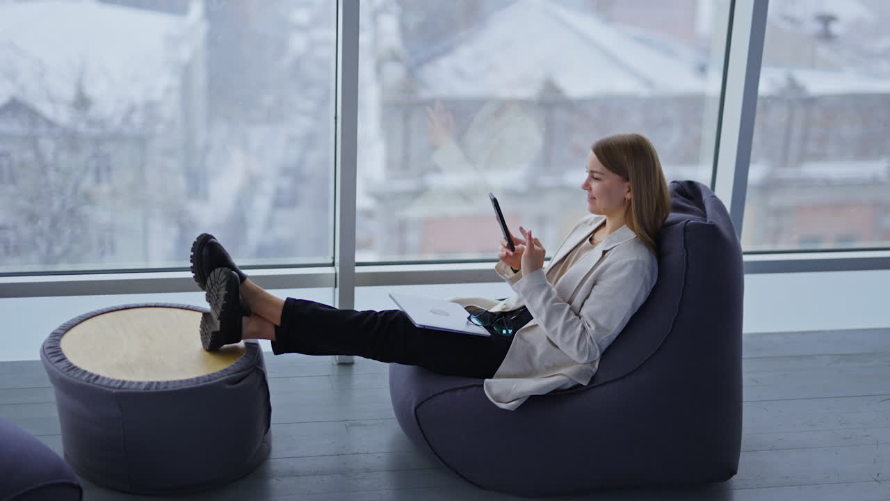 Relaxed young woman sits in bean chair stretching her legs to the coffee table. Lady with laptop and glasses on her knees looks at her phone and smiles. Snowing weather backdrop in blur.