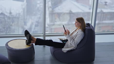 relaxed young woman sits in bean chair stretching her legs to the coffee table. lady with laptop and glasses on her knees looks at her phone and smiles. snowing weather backdrop in blur.