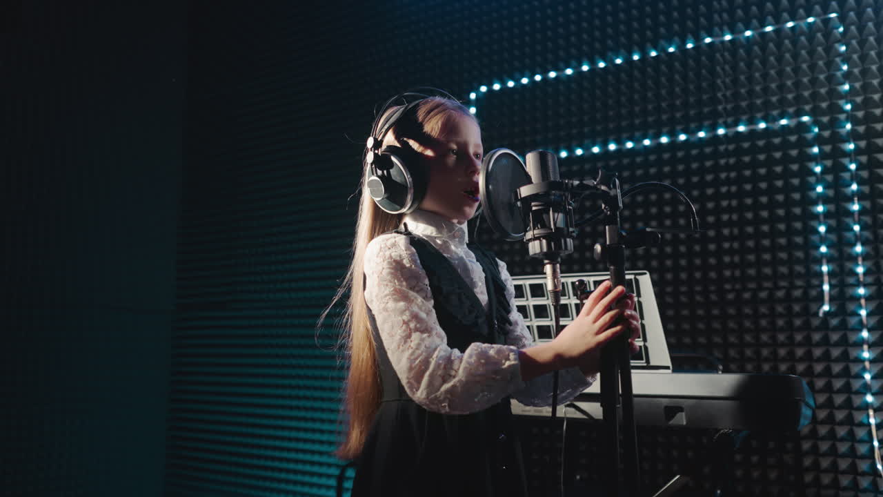 Child clings to microphone stand dancing in place. Little girl with long hair dances singing in microphone with headphones. Young artist records song