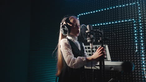 child clings to microphone stand dancing in place. little girl with long hair dances singing in microphone with headphones. young artist records song