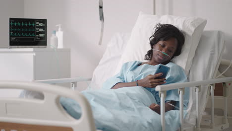 A-young-woman-writes-a-message-on-her-phone-while-lying-in-a-hospital-ward.-An-African-girl-is-lying-in-a-ward-connected-to-ECG-and-oxygen-devices-in-a-mask-and-writes-messages-to-relatives