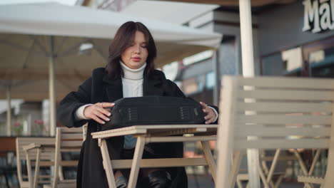 lady seated outdoors at mall unzips bag on wooden table in modern urban setting, stylish outfit includes black coat and white sweater, background features tables, chairs, and blurred storefronts