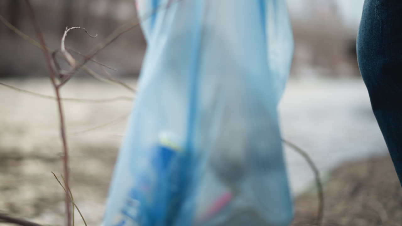 Close-up of volunteer wearing orange gloves picking up dirty plastic bottle from muddy ground with dry twigs, placing it into blue bag during environmental cleanup to reduce pollution