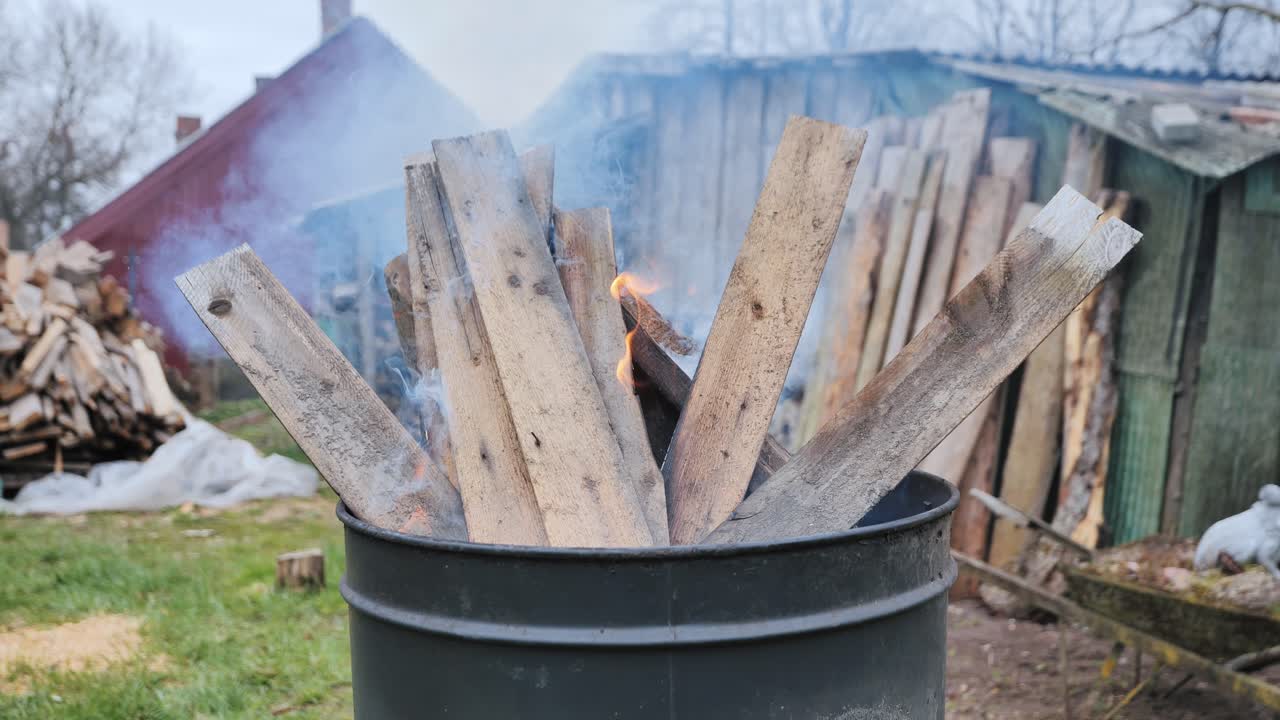 Smoke rising from wooden planks burning in metal barrel in rustic Latvian yard