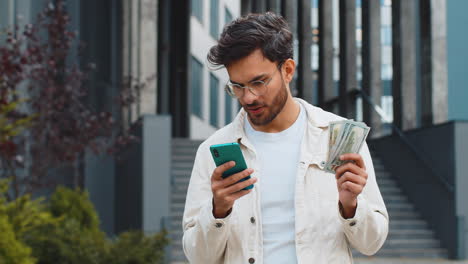happy indian man counting money dollar cash use smartphone calculator app in urban city street