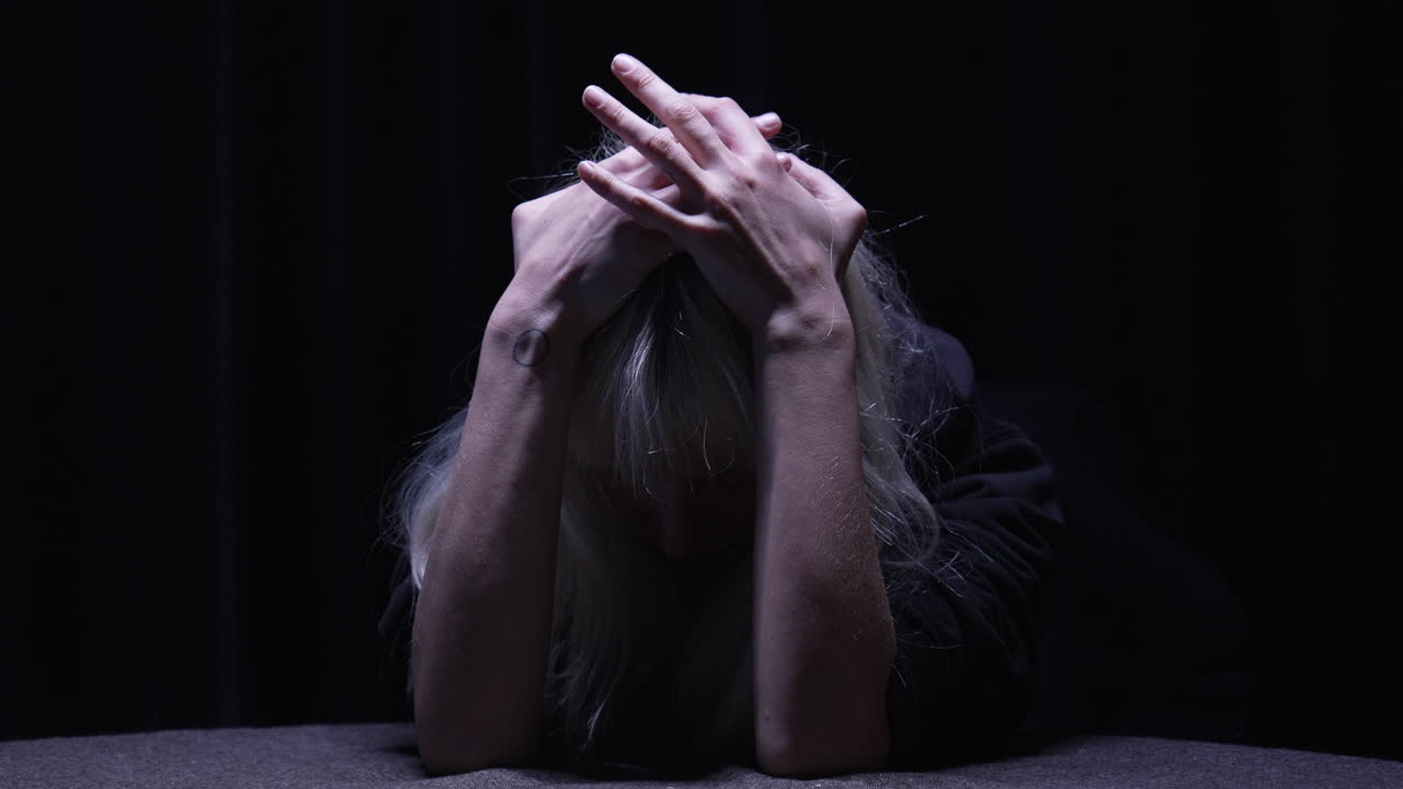 불안한 사람이 손으로 머리를 감싸고 책상에 앉아 머리 위 조명 하나가 비추는 어두운 방에서 손가락이 초조하게 안절부절합니다.