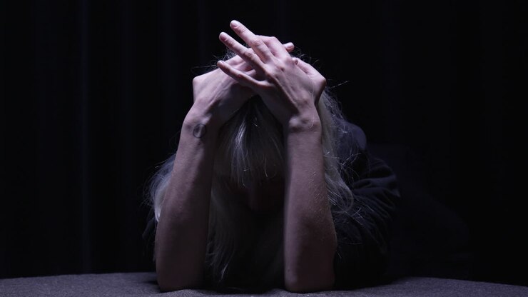 Persona ansiosa seduta a una scrivania con la testa tra le mani, le dita che giocherellano nervosamente in una stanza scarsamente illuminata da un'unica luce dall'alto