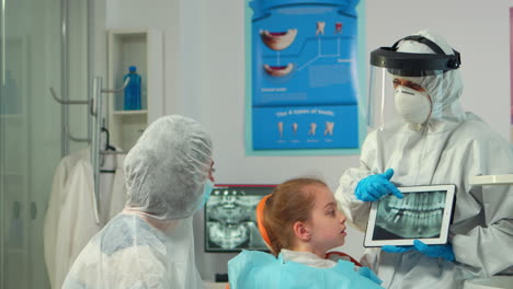 dentist in protective equipment showing on tablet dental x-ray of child