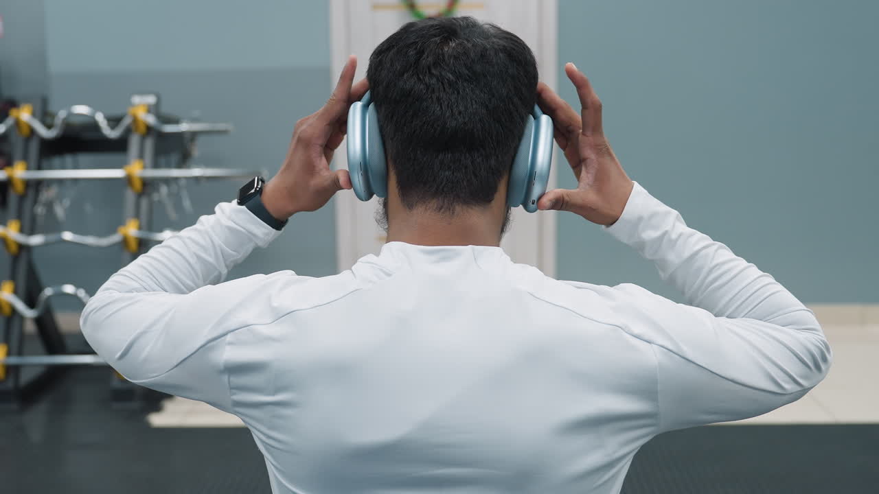 Rear view of athlete adjusting headphones while preparing to lie on bench press, focused on lifting weight, barbell on rack behind, gym environment with steel bar, window view, building in background