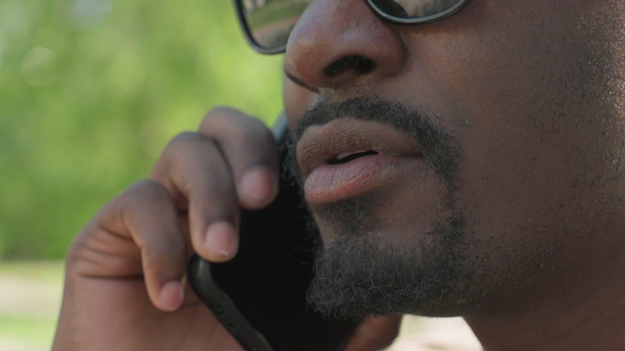 Close up of man wearing glasses holding and using smartphone outdoors with greenery in background. subject facial features including beard texture and skin details are visible as man