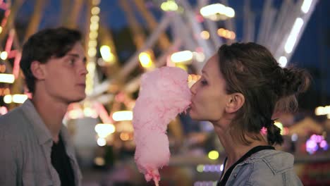 lovely young couple at amusement park. spending time together, eating pink cotton candy from sides. loving couple, dating. blurred funfair view on the background