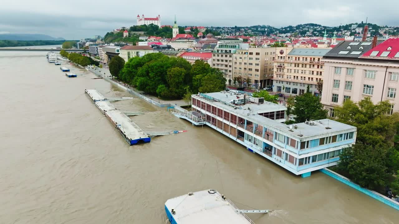 Aerial footage shows the flooded riverfront in Bratislava, where rising waters from the Danube River have submerged parts of the embankment. Prominent buildings and boats line the waterlogged area.