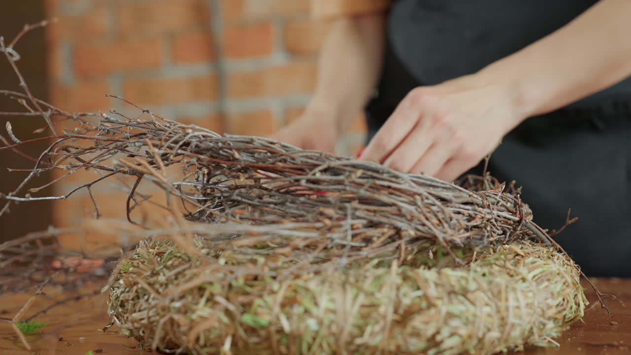 Female florist hands creating handmade wreath by arranging dry twigs on straw base, focusing on natural craft process with rustic materials on wooden table for decoration or seasonal design project