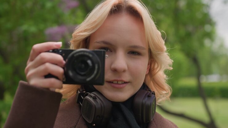 Shows woman outdoors wearing brown coat and black scarf. Woman holds digital camera up to take photos with headphones around her neck set against green trees and purple flowers in park