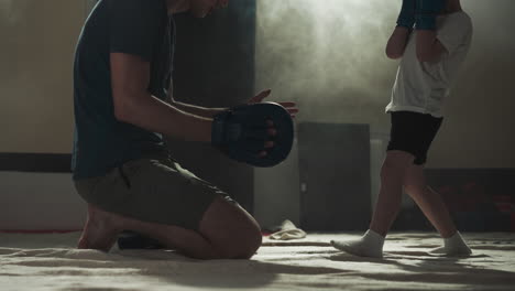 child kicks pad on instructor hand in fighting gym. caring trainer teaches toddler boy to hit by legs in sports center slow motion. kickboxing lesson