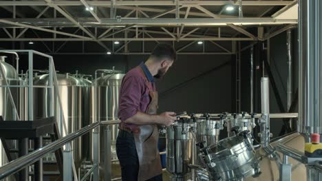 young male brewer wearing a leather apron supervise the process of beer fermentation at a modern brewery factory