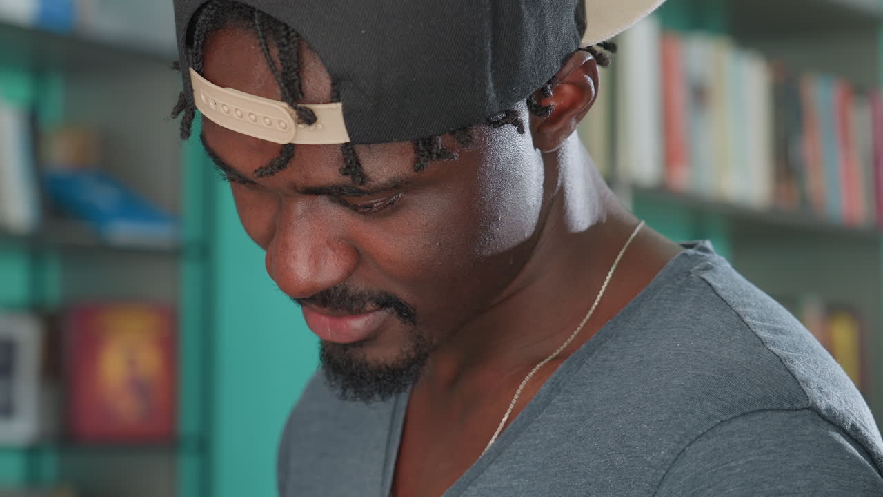 Close up of young man wearing backward cap smiling gently while looking down with focused expression, in softly lit indoor setting with teal background