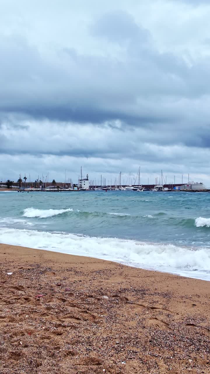 Rekaman panning vertikal Pantai San Sebastian yang kosong dengan ombak bergulir di bawah langit mendung, Barcelona, Spanyol