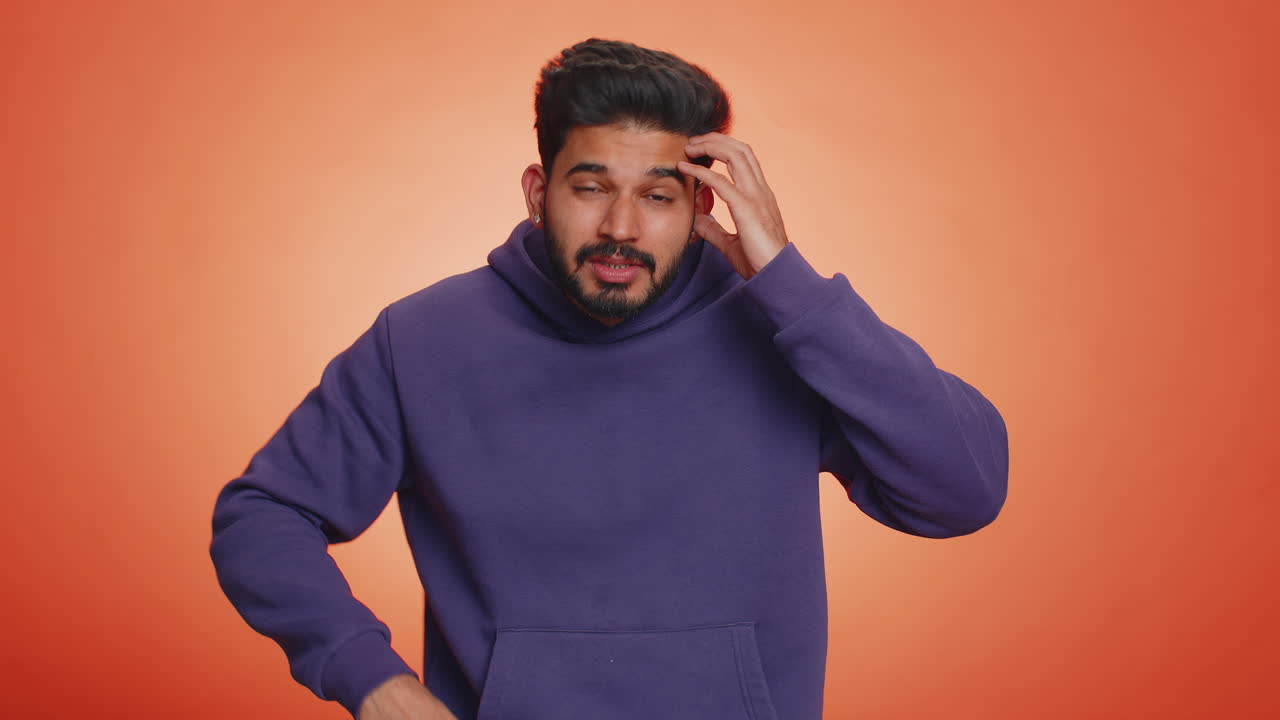 A young man in a purple hoodie is standing in front of an orange background. He is looking at the camera with a confused expression and is making a hand gesture as if asking a question.