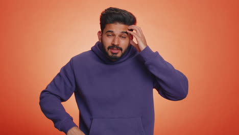 un joven con una sudadera púrpura está de pie frente a un fondo naranja. está mirando a la cámara con una expresión confusa y está haciendo un gesto de mano como si estuviera haciendo una pregunta.