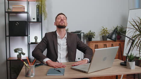 Businessman-working-on-laptop,-meditating,-doing-yoga-breathing-exercise-in-lotus-position-at-office