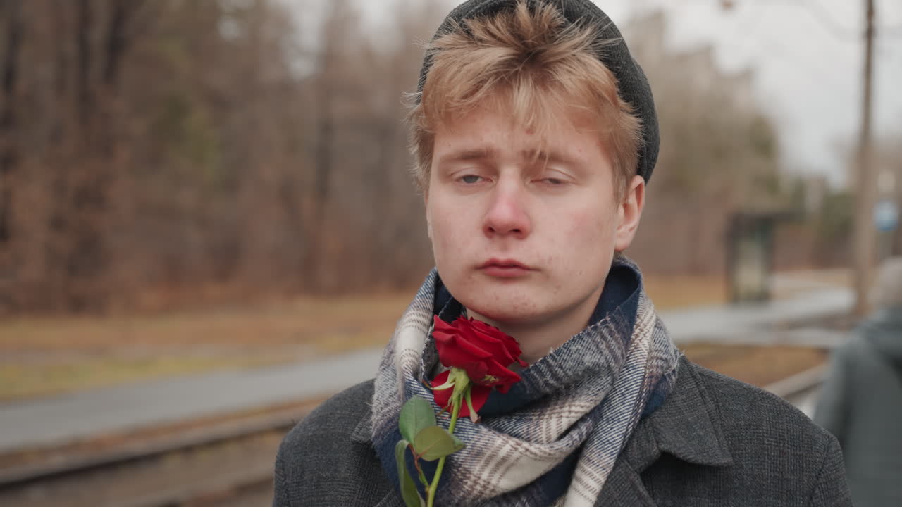 Young white man holding rose at train platform, poet creator waiting in cold autumn, wool scarf and beanie, coat, soft cinematic portrait, melancholic gaze, muted color palette, shallow focus, lonely