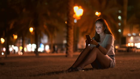 Ein-Schönes-Junges-Mädchen-Im-Sommer-In-Einer-Großen-Nachtstadt-Hält-Ein-Smartphone-In-Der-Hülle-Und-Schreibt-Eine-Nachricht,-Liest-E-Mails-Und-Kommuniziert-In-Chatrooms-Und-Sozialen-Netzwerken,-Führt-Einen-Blog-Und-Schaut-Auf-Den-Telefonbildschirm