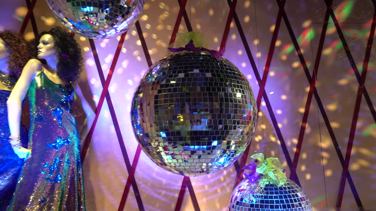 A showcase is decorated with two women mannequins are dressed at a shiny holiday dresses and a disco ball with shiny rays and purple light