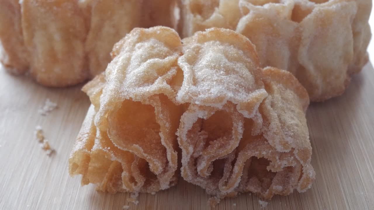 Zoom out reveals a golden Spanish sweet, Flor Frita, resting on a wooden board dusted with sugar