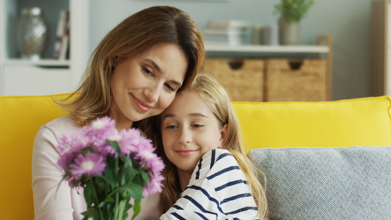 Clore Up View Of Mother Hugging Her Cute Teenager Daughter Sitting On Sofa While Holding A Bouquet Of Flowers