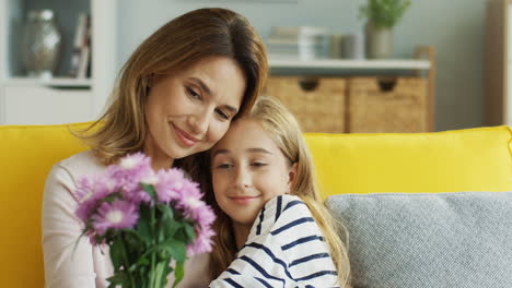 clore up view of mother hugging her cute teenager daughter sitting on sofa while holding a bouquet of flowers