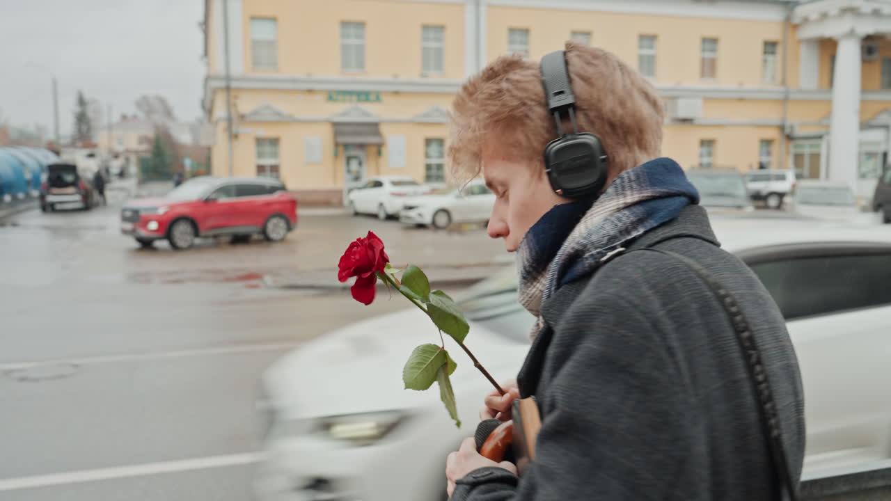 Romantický básník s květinou. Městská scéna s deštěm a sluchátky. Zranitelný mladík s růží na deštivé městské ulici. Mladý běloch nesoucí květinu, zatímco déšť padá na rušný městský chodník