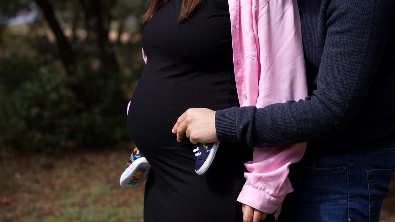 Pregnant couple holds tiny baby shoes near belly in forest outdoors with copy space on left side