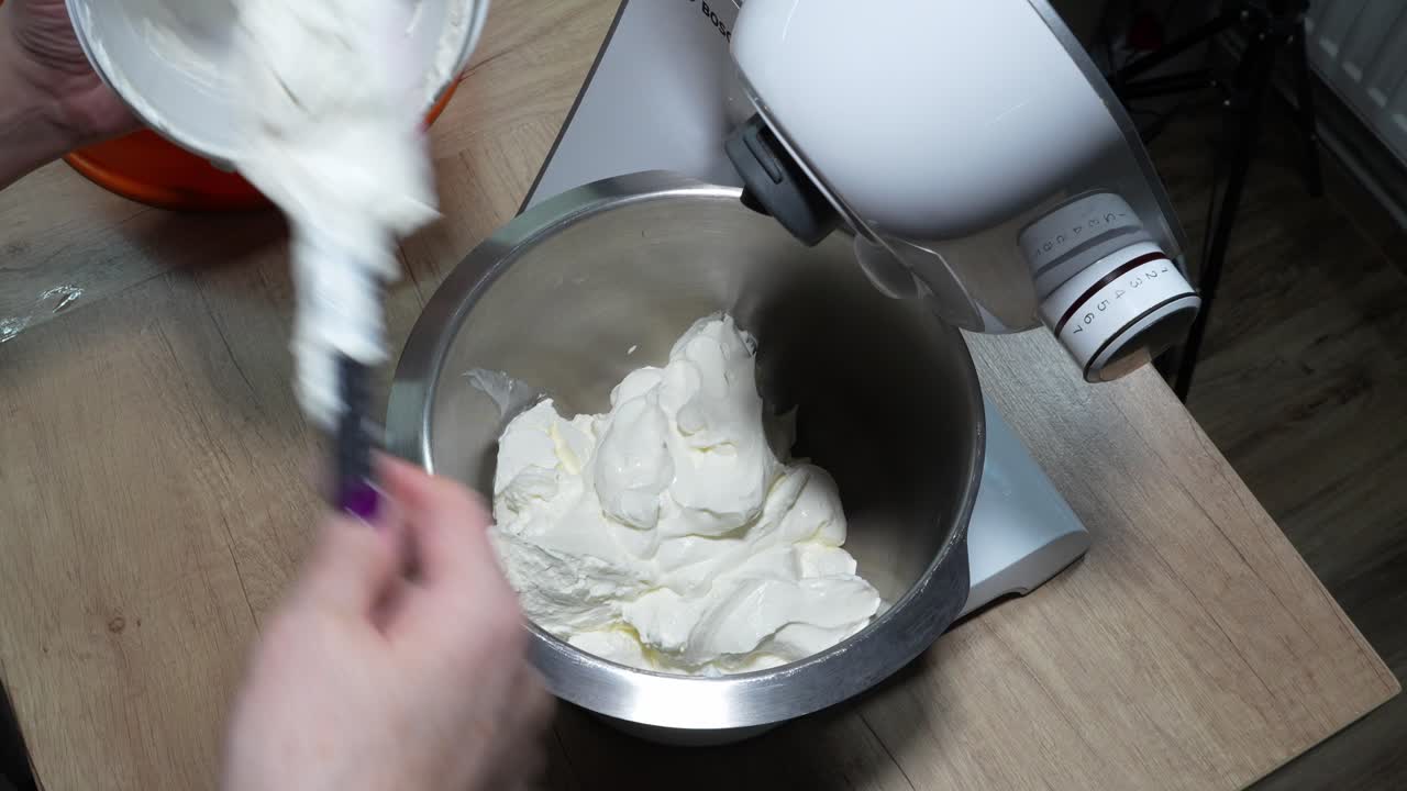 A close-up view of hands placing cottage cheese into a stand mixer bowl in a kitchen setting.
