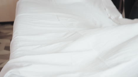 fair skin woman dressed in white trouser adjusts smooth white silk sheet over light gray bed, partially visible in soft natural light, emphasizing texture and gentle movement in clean setting