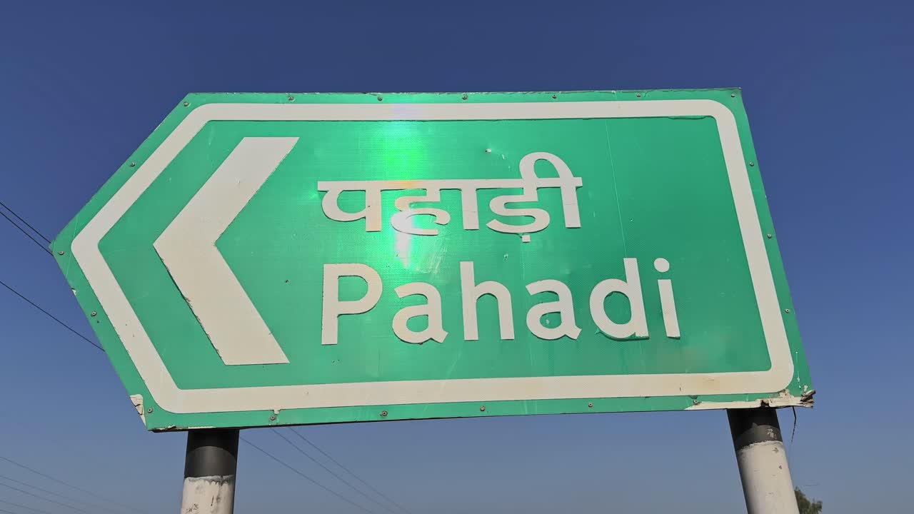 Primo piano di un cartello stradale verde con la scritta Pahadi in hindi e in inglese con freccia a sinistra, montato su pali di metallo con cielo azzurro sullo sfondo in pieno giorno