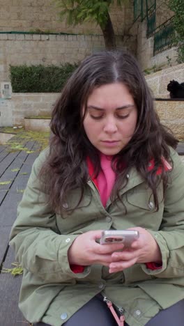 young adult woman using her smartphone while sitting on a bench outdoors