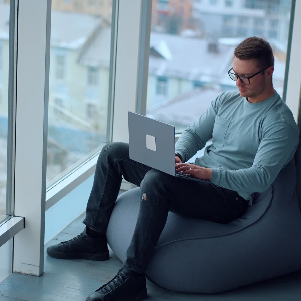 Young man sitting comfortably in bean bag chair working on his laptop. Busy man works near the big window. View from top