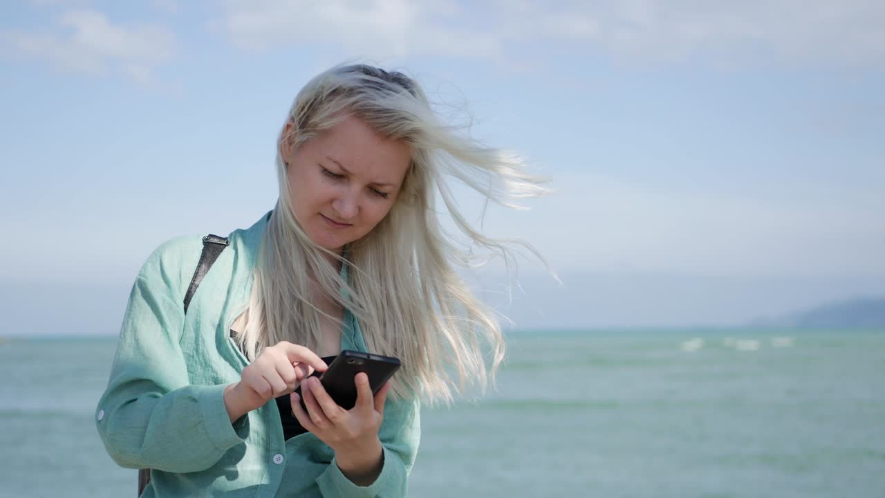 Beautiful slim woman with long blonde hair and green shirt standing and using smartphone over background sea. Girl touching screen