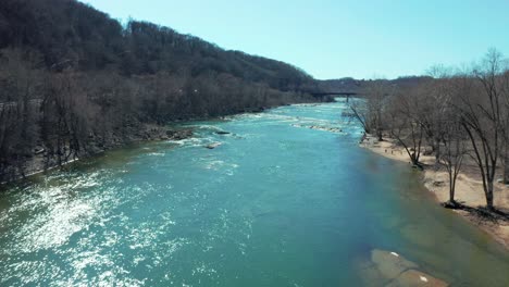 aerial dolly of a river cutting through a mountainous landscape, with sparkling waters and leaveless trees lining the riverbanks