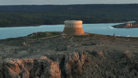 drone flying over defence tower in menorca spain to reveal small township of fornells at sunset