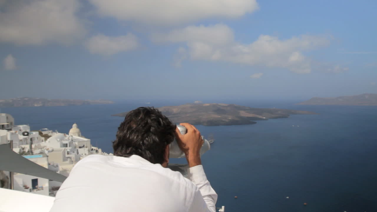un hombre vestido con una camisa blanca se acerca a un par de binoculares públicos que supervisan la vista de la caldera de santorini, incluida la isla volcánica de nea kameni