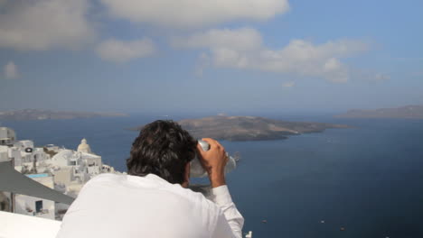 a man wearing a white shirt approaches a pair of public binoculars that oversee the view of the santorini caldera including the volcanic island of nea kameni