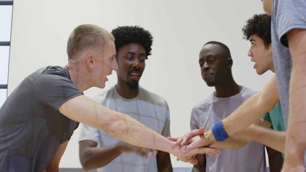 Diverse male team forming huddle in gym left player holding volleyball urging play stacking hands