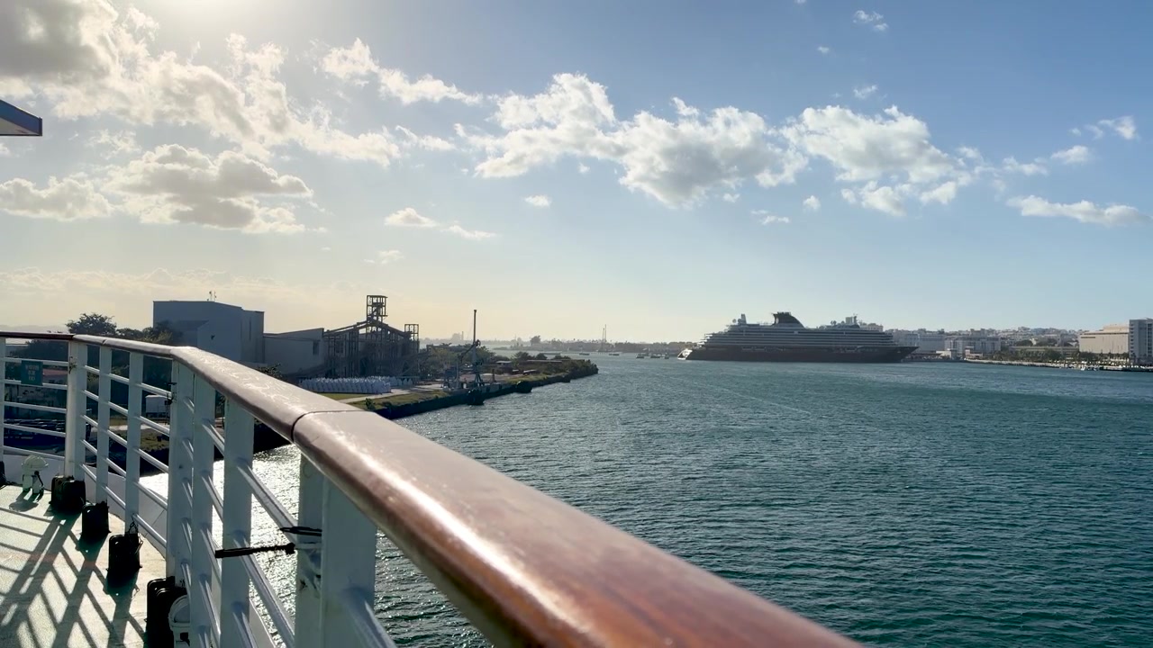 POV-Aufnahme vom Deck eines Kreuzfahrtschiffes mit Holzgeländer, mit Blick auf einen sonnigen karibischen Hafen mit industriellen Hafenanlagen und einem fernen Schiff.