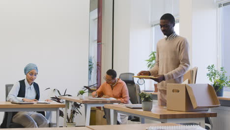 Muslim-Woman-And-Sitting-At-A-Table-In-The-Office,-While-A-Young-Man-Takes-Out-His-Personal-Items-From-A-Box-And-Places-Them-On-The-Table-1