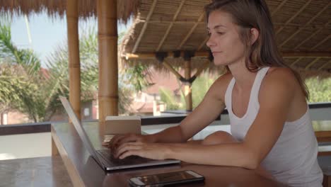 niña escribiendo en una laptop, respondiendo una llamada telefónica y escribiendo algo en la agenda mientras se sienta en un taburete en la playa