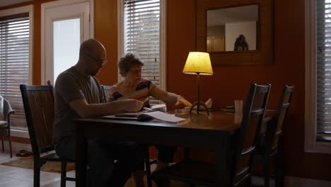 a middle-aged man, and senior woman sit together at a dining table in a warmly lit home, sorting through receipts. son is focused on counting and calculating receipts, onlooking mother instructs.