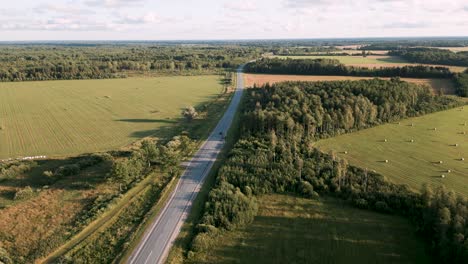 Vuelo-Ascendente-De-Drones-Sobre-Una-Carretera-Rural-En-Estonia,-Europa-Siguiendo-A-Un-Auto-Solitario-Conduciendo-A-Lo-Largo-De-La-Calle-Vacía-Entre-Campos-Y-Pequeños-Bosques-En-El-Verano-De-2022---Vista-De-Pájaro-Tomada-Como-Publicidad